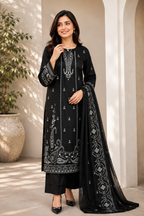 A woman models a black traditional Indian salwar kameez suit with intricate silver embroidery, paired with matching black trousers and a sheer black dupatta, set against a warm beige architectural background.